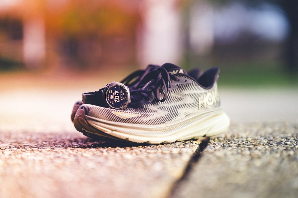 A pair of Hoka running shoes with a Garmin watch beside them on a sunny outdoor track.