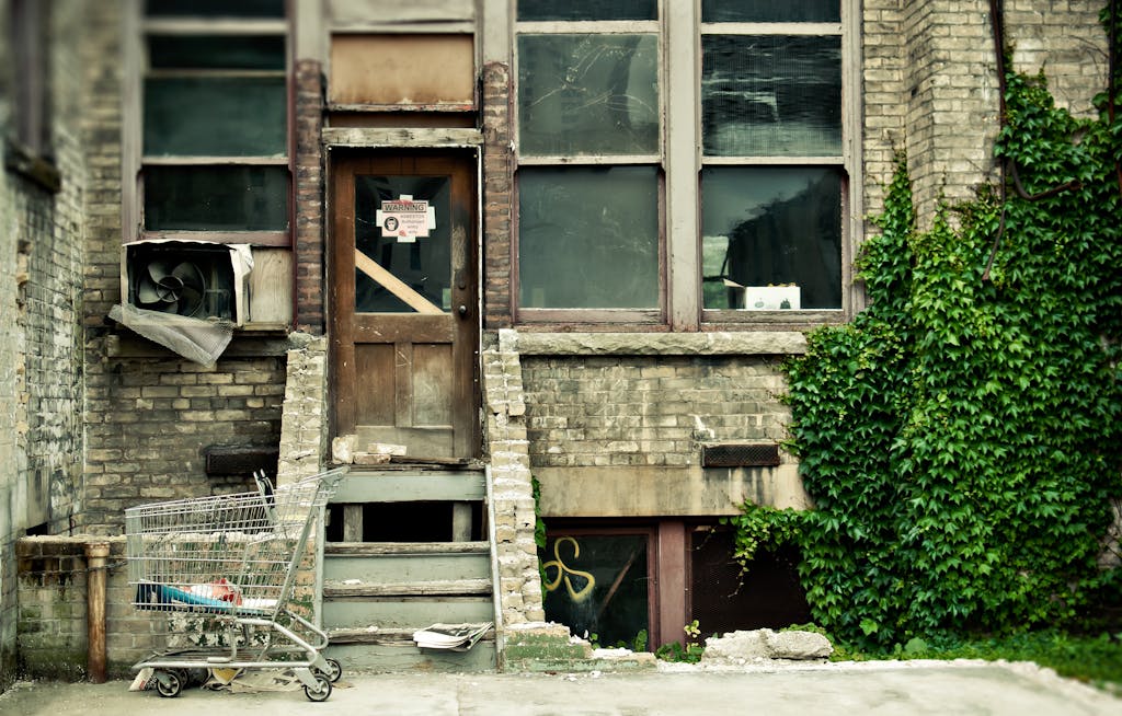An abandoned building entrance with a broken door, windows, and a shopping cart on the steps.