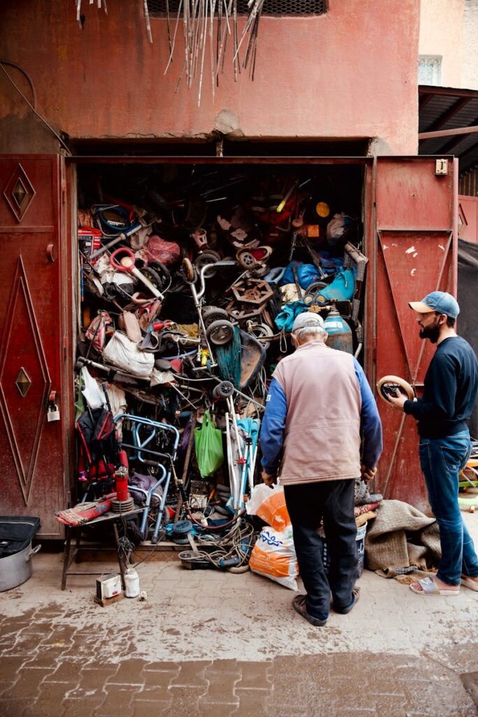 Two men inspecting a workshop with a cluttered collection of discarded items and tools.