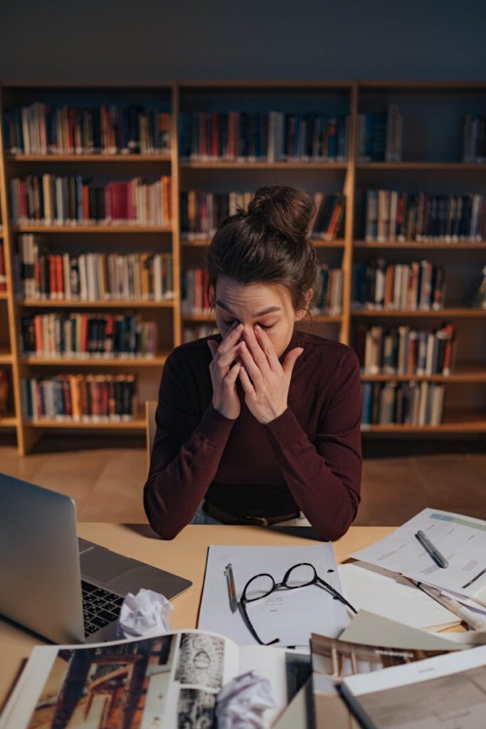Photo by Ron Lach Woman feeling stressed while studying in a library, surrounded by books and laptop.